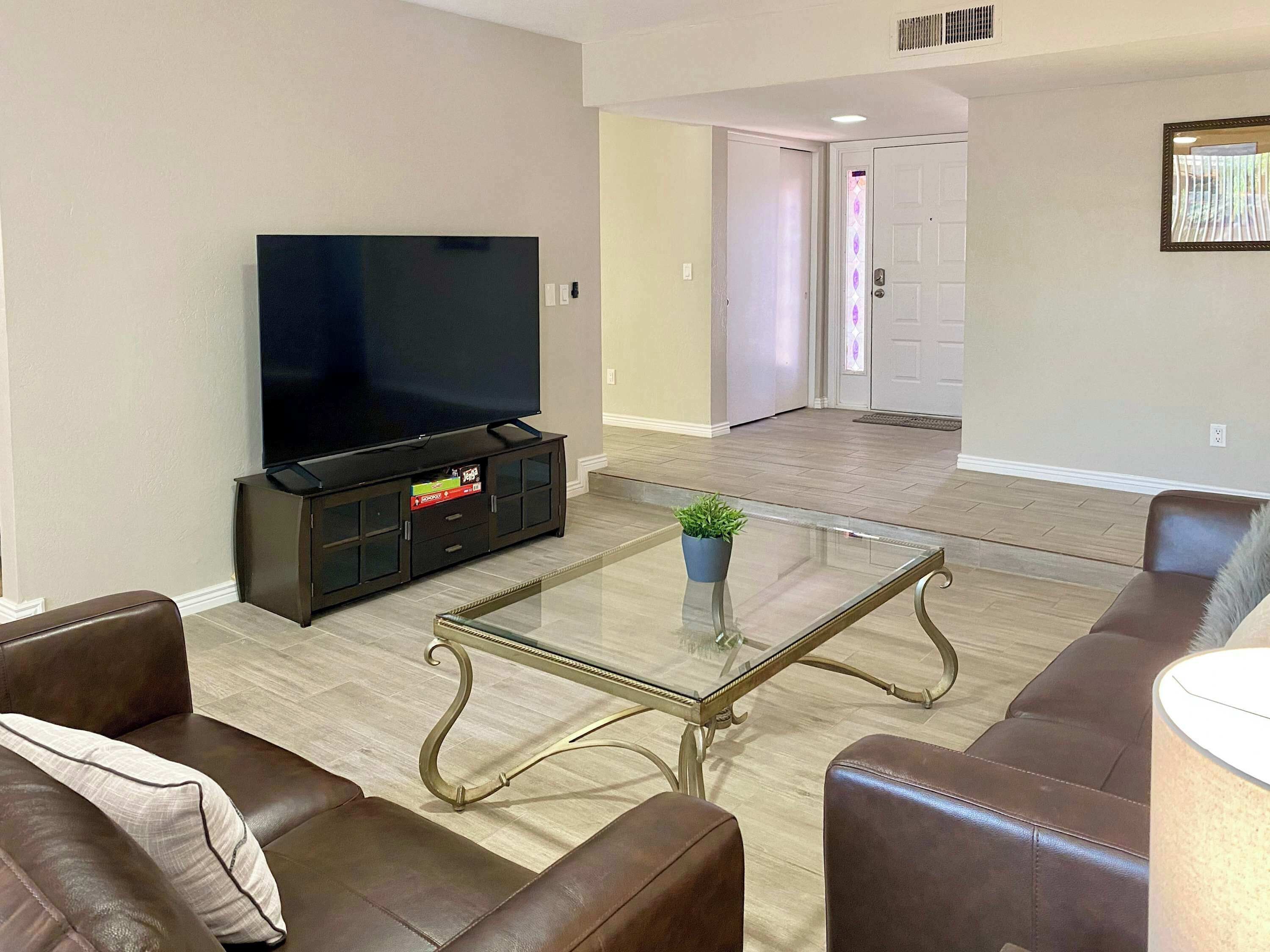A living room with a flat-screen TV on a cabinet, a glass coffee table, brown leather sofas, and a door in the background.