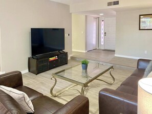 Un salón con una televisión de pantalla plana sobre un mueble bajo, una mesa de centro de vidrio, sofás de cuero marrón y una puerta al fondo.