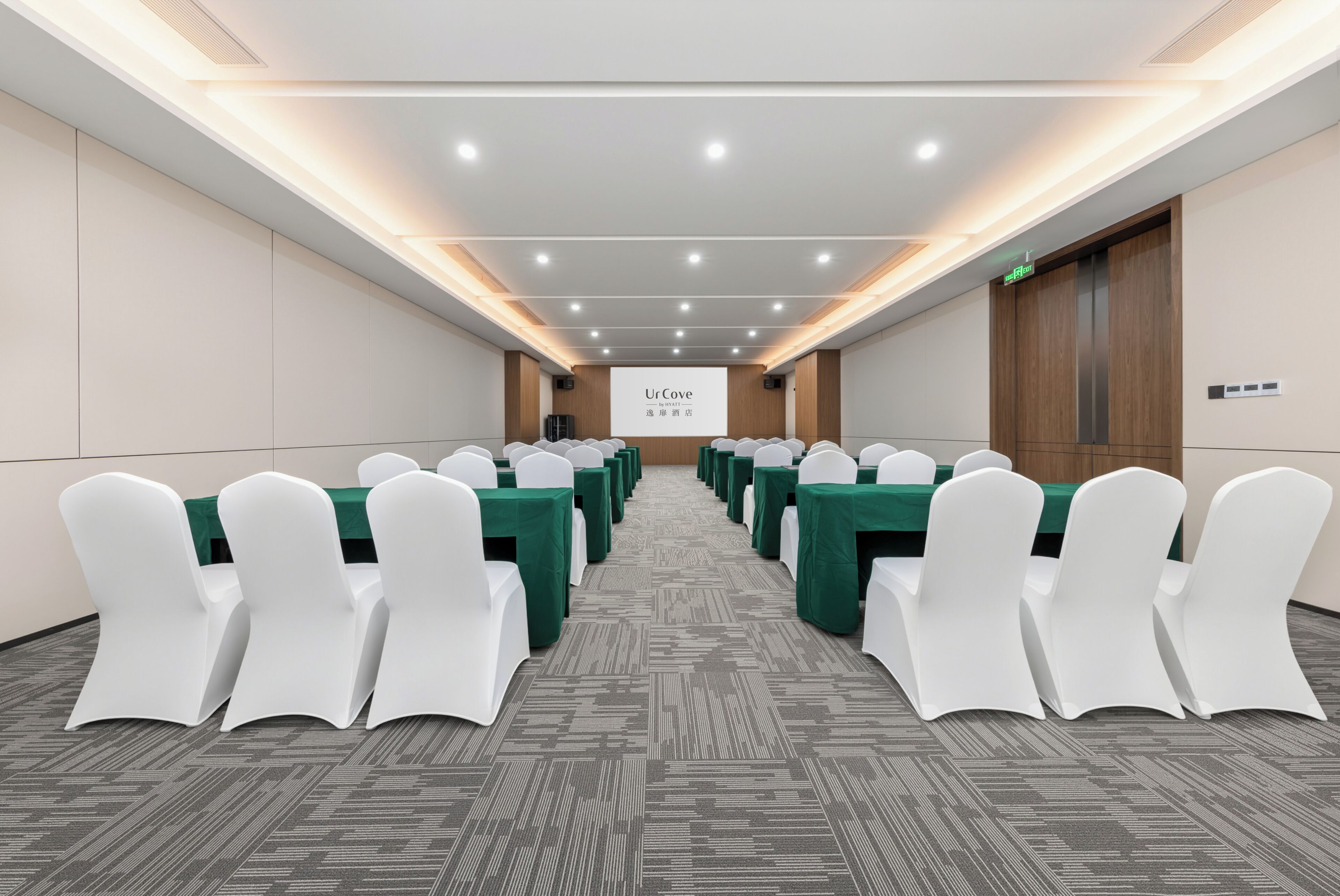 Una sala de conferencias con filas de sillas blancas y mesas verdes, una pantalla de proyector y una puerta de madera.