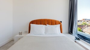 Ein Schlafzimmer mit einem Bett, weißer Bettwäsche, einem orangefarbenen Kopfteil, einem Nachttisch, einer Taschentuchbox und einem Fenster mit Blick auf Gebäude.