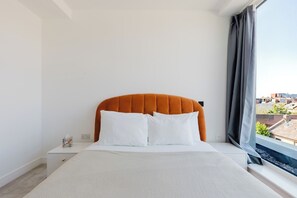 Ein Schlafzimmer mit einem Bett, weißer Bettwäsche, einem orangefarbenen Kopfteil, einem Nachttisch, einer Taschentuchbox und einem Fenster mit Blick auf Gebäude.