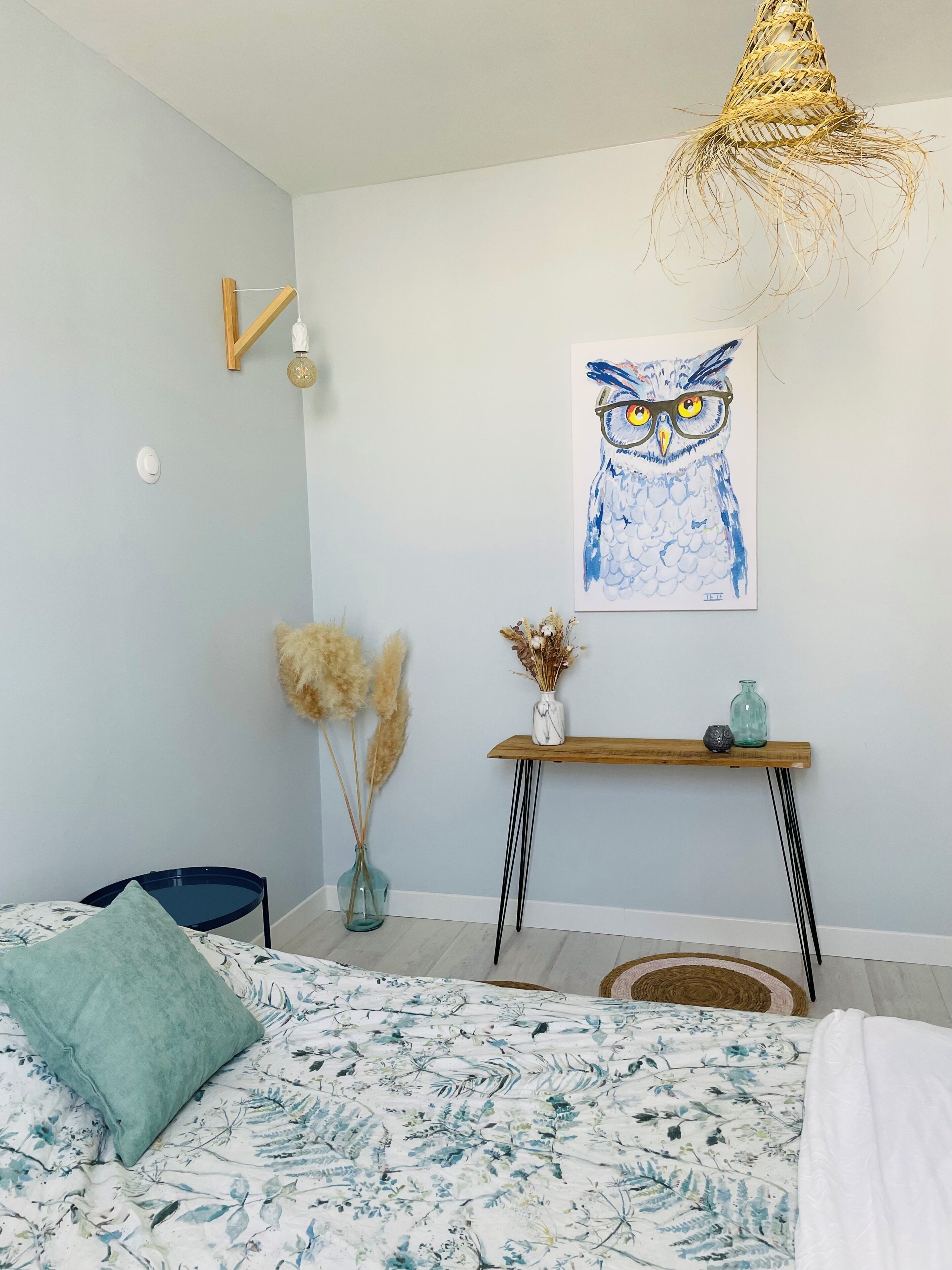 Quarto com cama, mesa de cabeceira com vaso, luminária na parede e pintura de uma coruja.