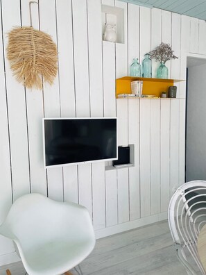 Ein modernes Wohnzimmer mit einem an der Wand befestigten Fernseher, weißen Möbeln, einer Hängeleuchte aus Weidengeflecht und dekorativen Objekten auf Regalen.