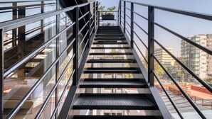 Eine Metalltreppe mit Glasgeländern führt zu einem Außenbereich, im Hintergrund sind Gebäude zu sehen.