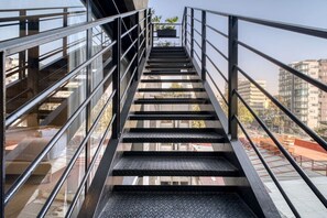 Eine Metalltreppe mit Glasgeländern führt zu einem Außenbereich, im Hintergrund sind Gebäude zu sehen.