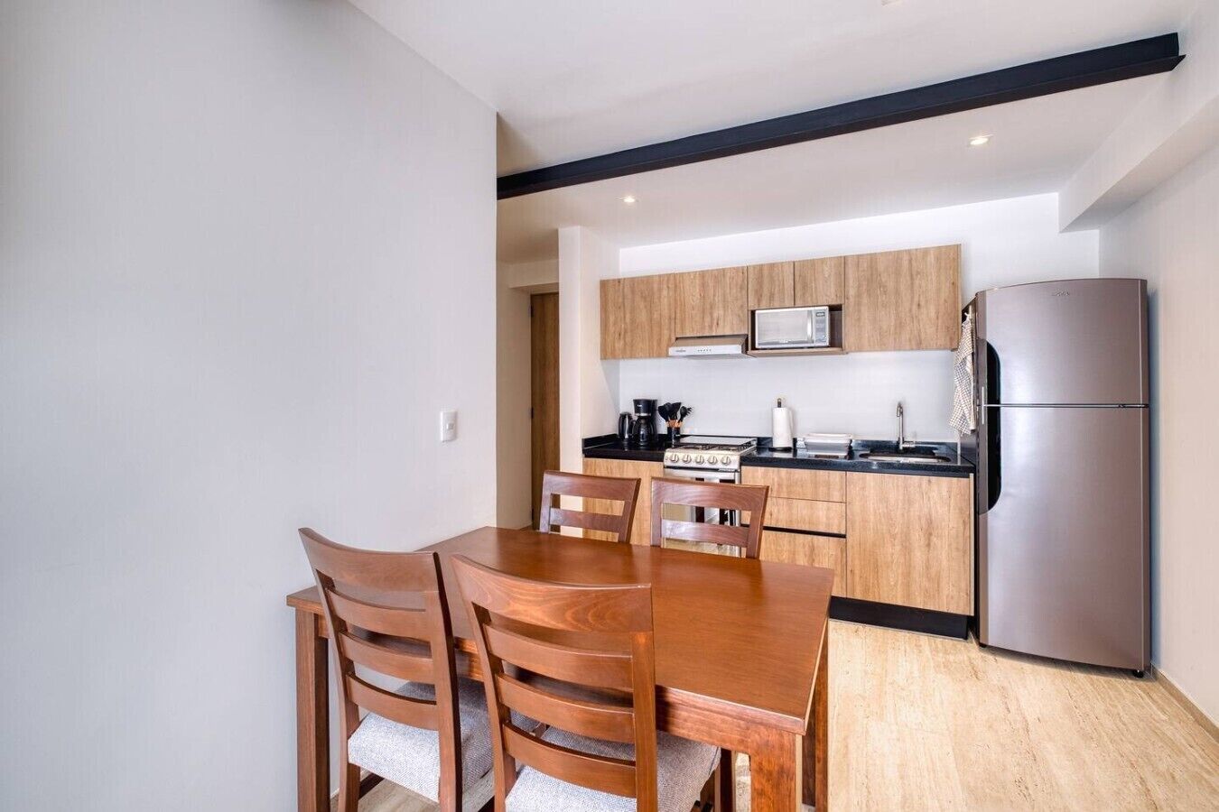 Een kleine keuken met houten kastjes, een roestvrijstalen koelkast, een magnetron en een eettafel met stoelen.