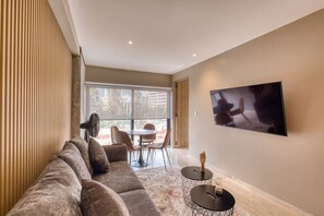 Ein modernes Wohnzimmer mit einer Couch, einem Esstisch mit Stühlen, einem an der Wand befestigten Fernseher und einem Ventilator.