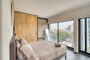 A modern bedroom with a large bed, a wooden wardrobe, and a city view through large windows. - Myla 904C - 1BR In Roma Norte - HighF (Mexico City)