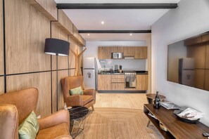 Ein modernes Wohnzimmer mit einer Couch, einem Couchtisch und einem Fernseher. Im Hintergrund ist eine Küchenzeile mit einer Mikrowelle, einem Herd und einem Kühlschrank zu sehen.