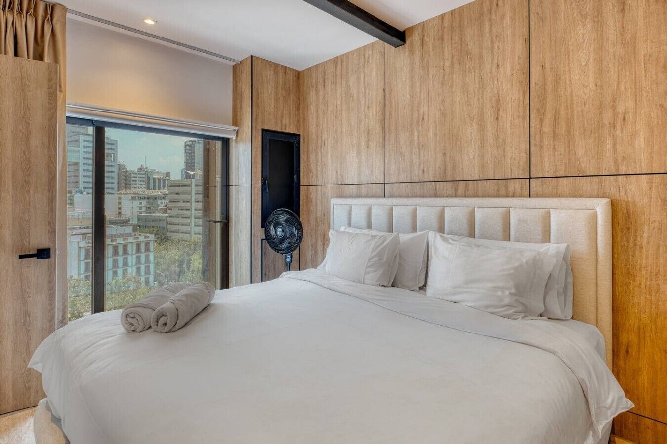 A modern bedroom with a large bed, a fan, and a view of the city through a large window.