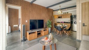 En moderne stue med et fladskærms-tv, træpaneler, et spiseområde med bord og stole, og et køkken i baggrunden.