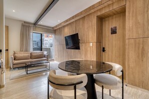 Una sala moderna con un sofá, una mesa redonda con sillas, un televisor de pantalla plana montado en la pared y un ventanal con cortinas.
