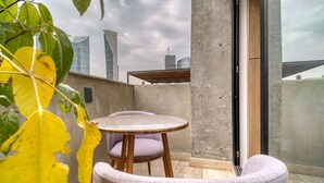 Ein Balkon mit rundem Tisch und zwei Stühlen, einer Betonwand und Blick auf Stadtgebäude.