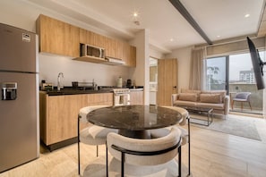 Eine moderne Küche mit Holzausbau, einer eingebauten Mikrowelle und einem Herd. Ein runder Esstisch mit zwei Stühlen steht daneben. Das Wohnzimmer bietet ein beiges Sofa und einen Flachbildfernseher.