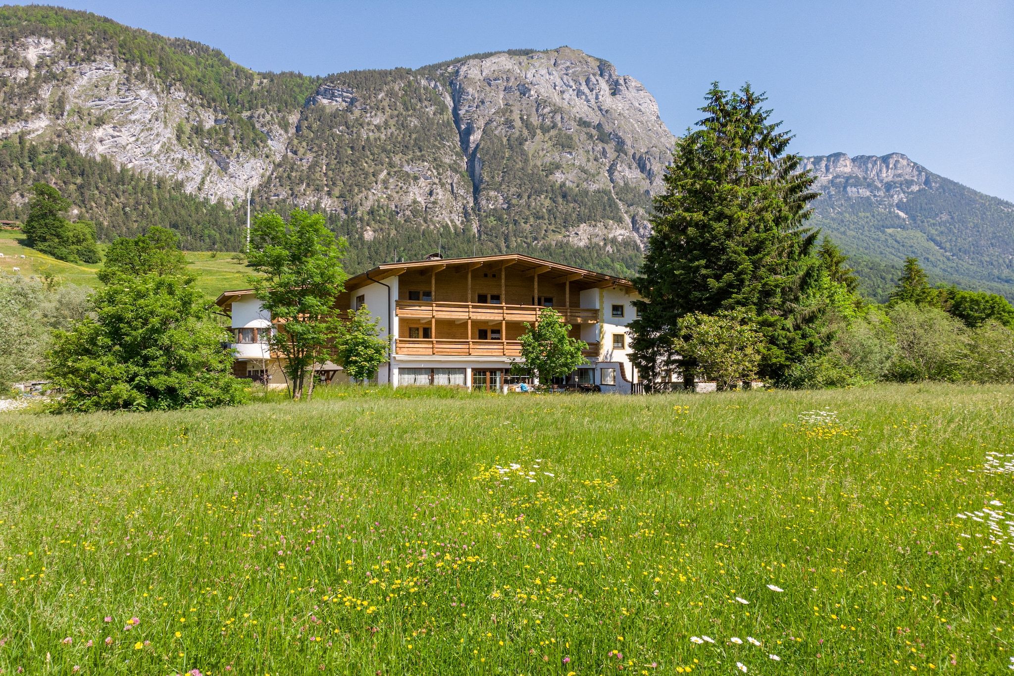 A two-story chalet with a balcony, surrounded by a grassy field and mountains in the background.
