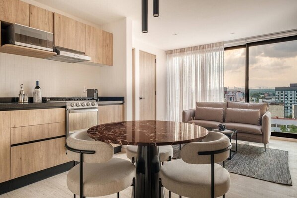 A modern kitchen with a built-in stove, a dining area with a round table and four chairs, and a large window with a city view. - Myla 04C - 2BR In Roma Norte - HighF (Mexico City)