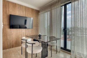 Ein modernes Wohnzimmer mit einem an einer Holzwand montierten Flachbildfernseher, einem Glastisch mit vier Stühlen und einem großen Fenster mit durchscheinenden Vorhängen.