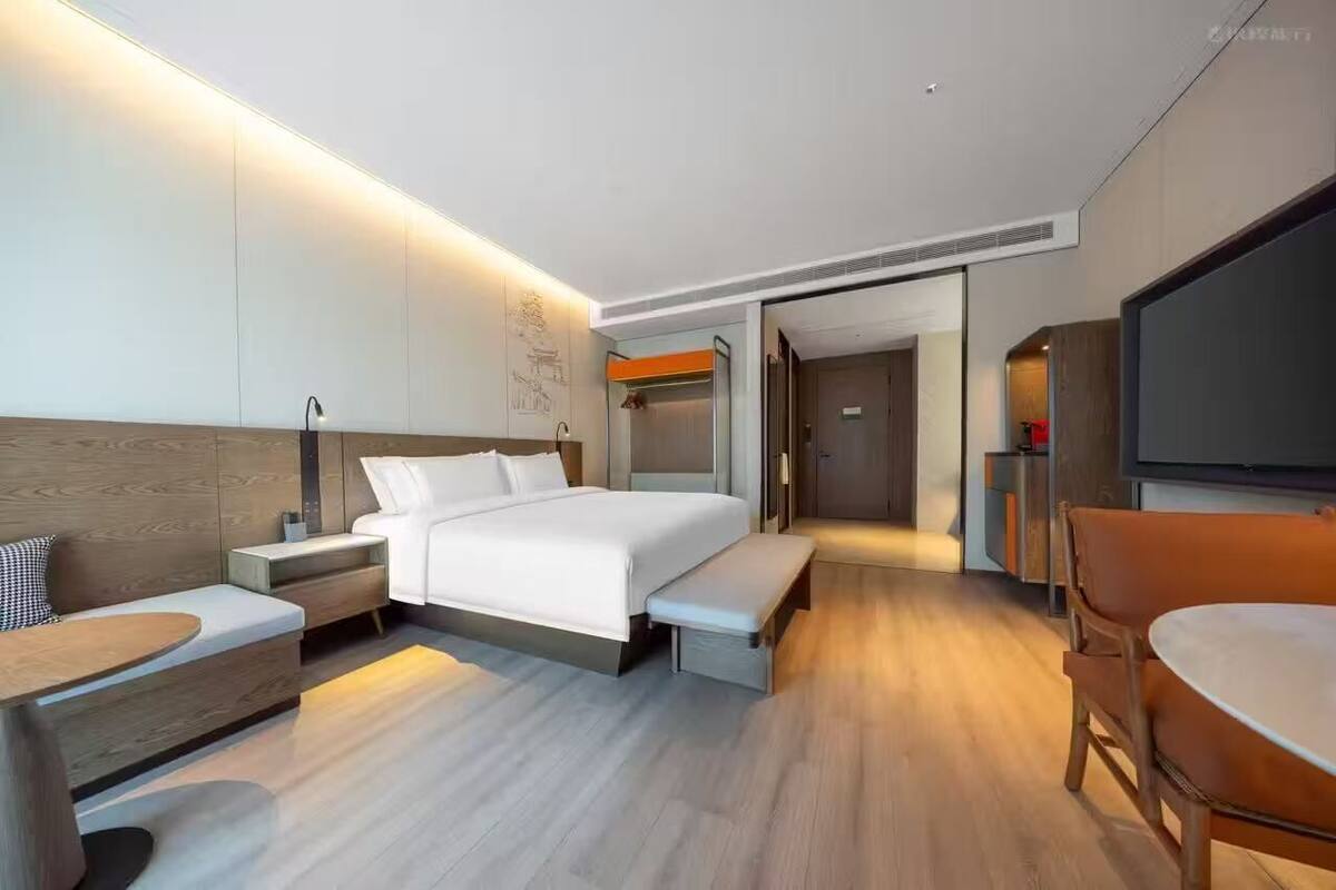 A modern hotel room with a bed, bedside tables, a bench, a TV, and a seating area.