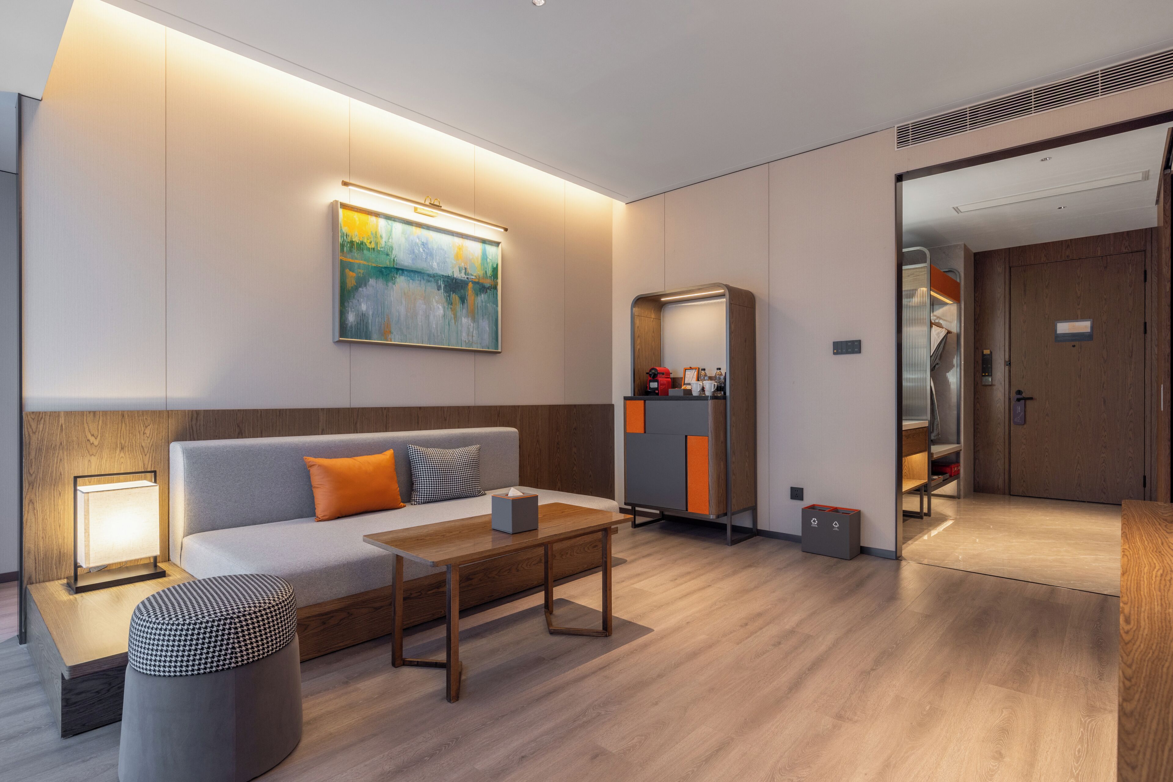 A modern hotel room with a sofa, a coffee table, a painting, a wardrobe, and a trash bin.
