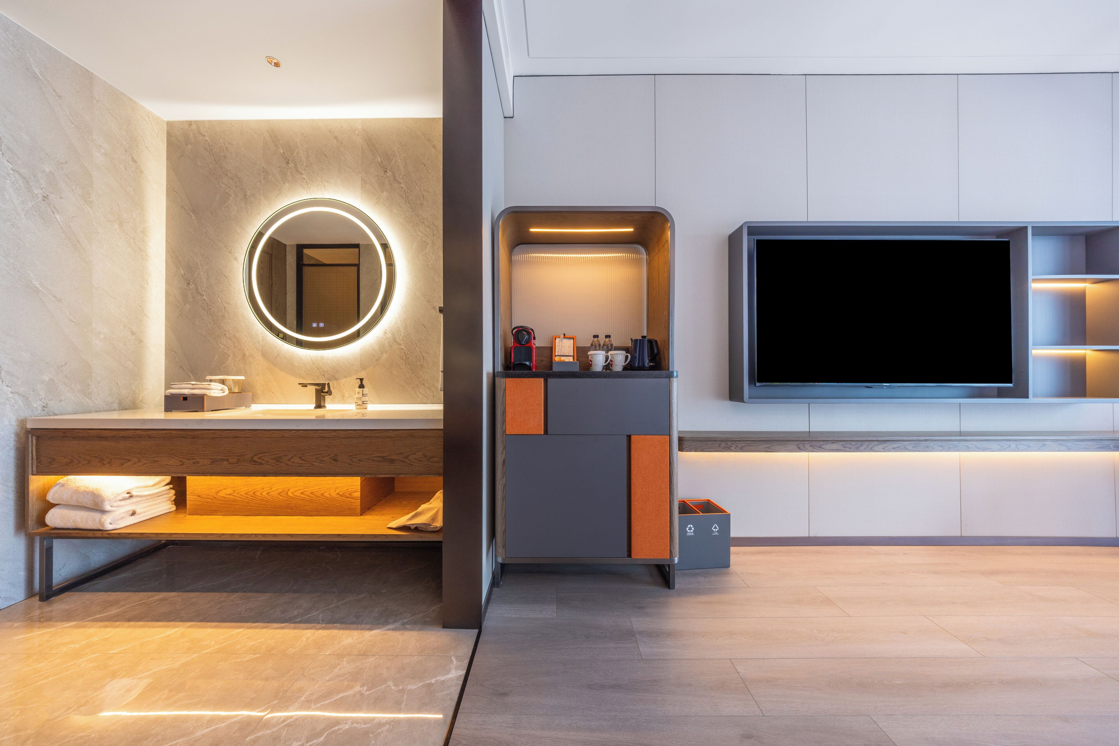 Une salle de bain moderne avec un meuble-lavabo, un grand miroir rond doté de lumières, un téléviseur et un petit coin salon.