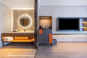 Een moderne badkamer met een wastafel, een ronde spiegel met verlichting, een televisie en een klein zithoekje.