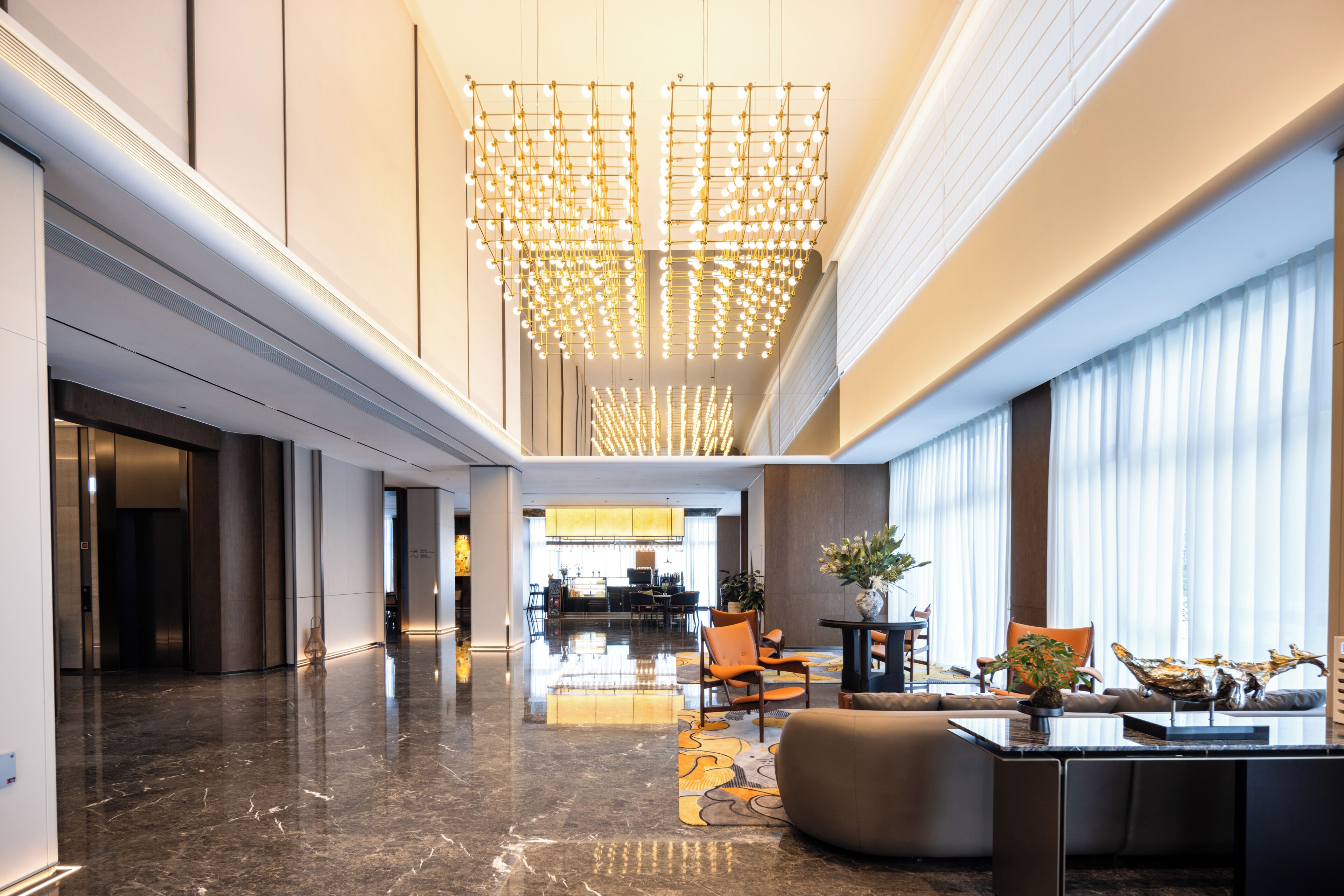 A modern hotel lobby with a central chandelier, seating area, and a reception desk.