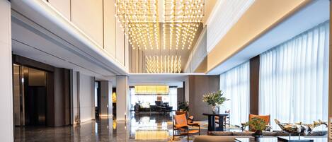A modern hotel lobby with a central chandelier, seating area, and a reception desk.