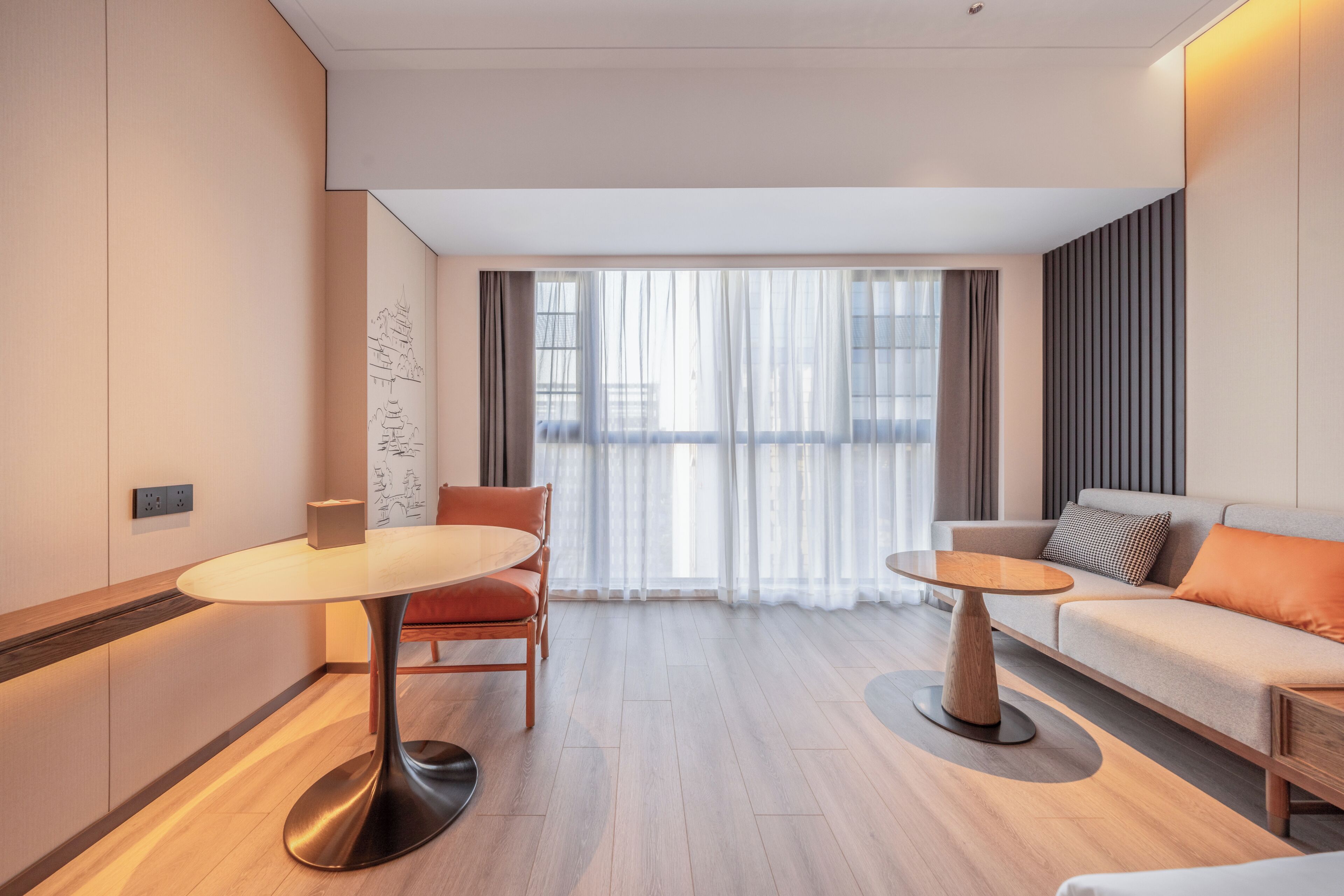 Een moderne hotelkamer met een sofa, twee tafels, een stoel en uitzicht op de stad door grote ramen.