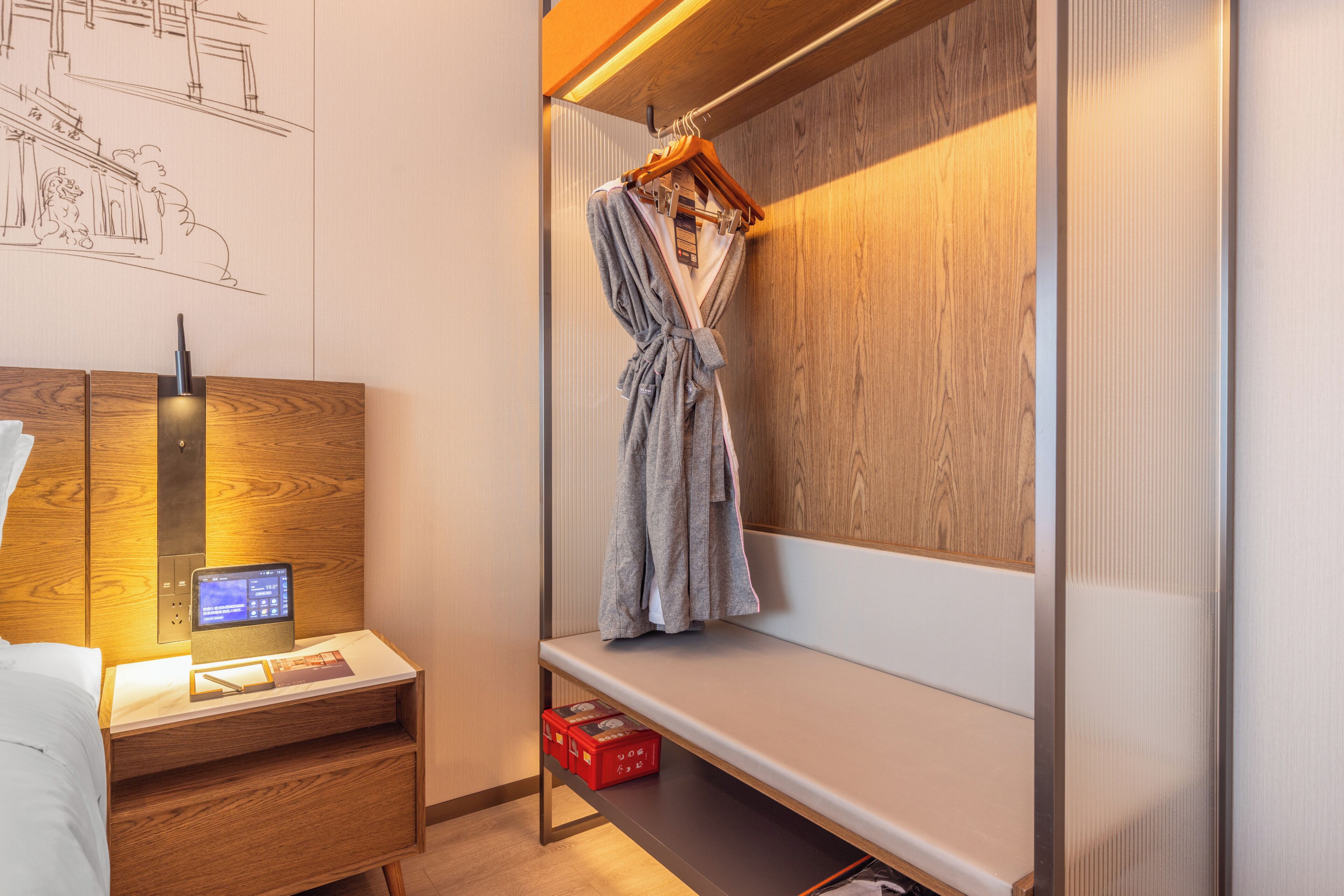 A modern hotel room with a wooden desk, a laptop, a hanging robe, and a shelf with red boxes.