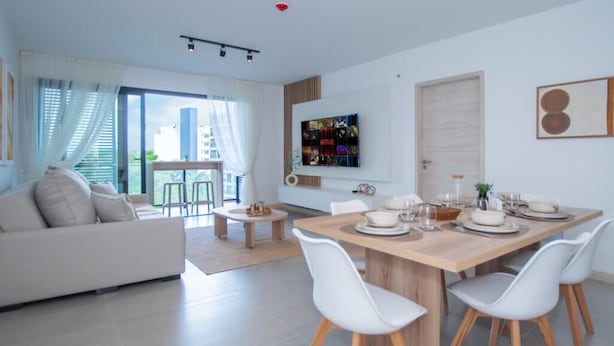 A modern living room with a sofa, dining table, and chairs. There is a TV mounted on the wall and a balcony with a view of buildings. - Corporate Stays Alban 1 (Asunción)