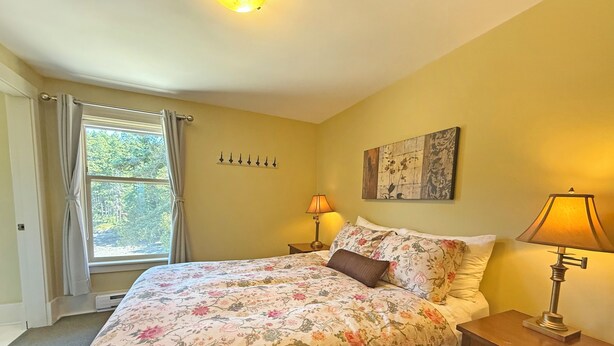 Une chambre à coucher comprenant un lit, deux lampes de chevet, une table de chevet, une fenêtre avec des rideaux et une œuvre d’art accrochée au mur.