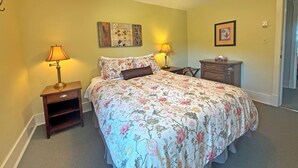 Une chambre à coucher avec un lit, deux lampes de chevet, une table de chevet, une commode, un tableau encadré au mur et une porte donnant sur une autre pièce.