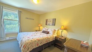 Une chambre à coucher comprenant un lit, deux lampes de chevet, une table de chevet, une commode en bois, une fenêtre avec des rideaux et une œuvre d’art accrochée au mur.