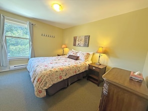 Un dormitorio con una cama, dos lámparas de noche, una mesita de noche, un aparador de madera, una ventana con cortinas y una obra de arte colgada en la pared.