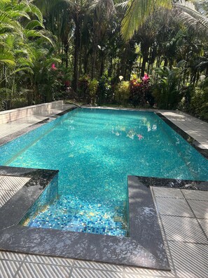 Une piscine rectangulaire à l’eau d’un bleu cristallin, entourée d’une terrasse carrelée et d’une végétation tropicale luxuriante.