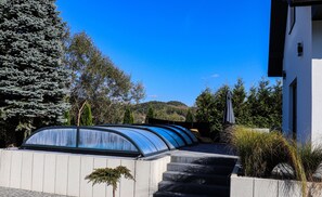 A modern building with a curved glass structure, steps leading to a door, and a view of a hill in the background.