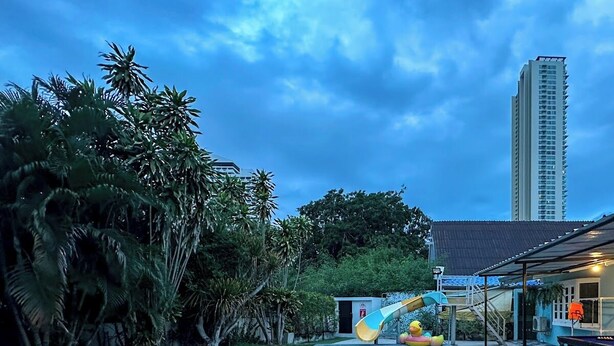 A swimming pool with a slide, surrounded by greenery and a tall building in the background. - MaMa Beach Pool Villa Pattaya (Muang Pattaya)