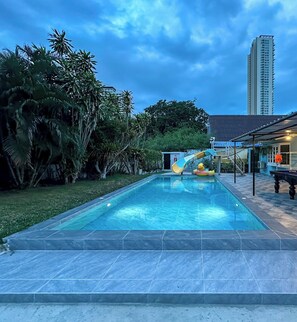 A swimming pool with a slide, surrounded by greenery and a tall building in the background. - MaMa Beach Pool Villa Pattaya (Muang Pattaya)