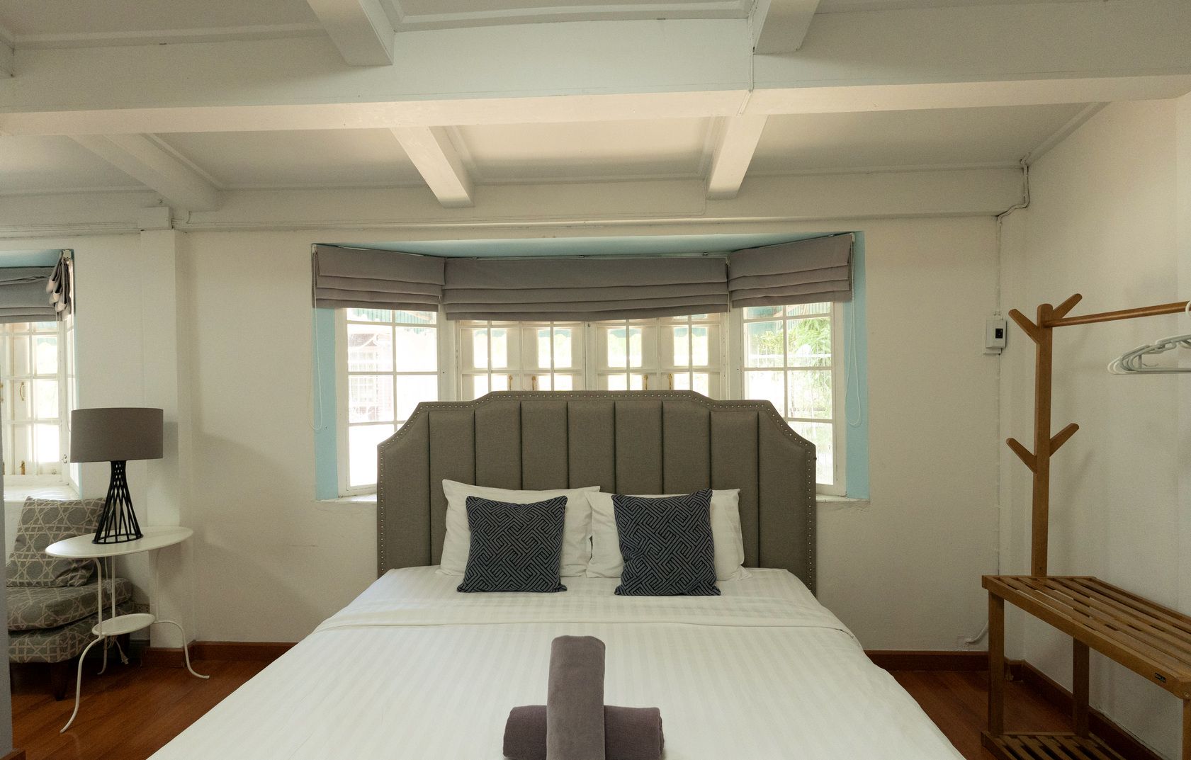 A bedroom with a large bed, a wooden coat rack, a small table with a lamp, and a window with curtains.