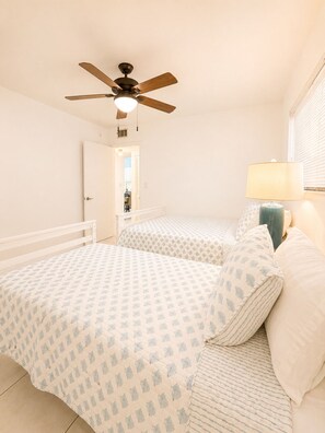 A bedroom with two beds, a ceiling fan, a nightstand with a lamp, and a door leading to another room.