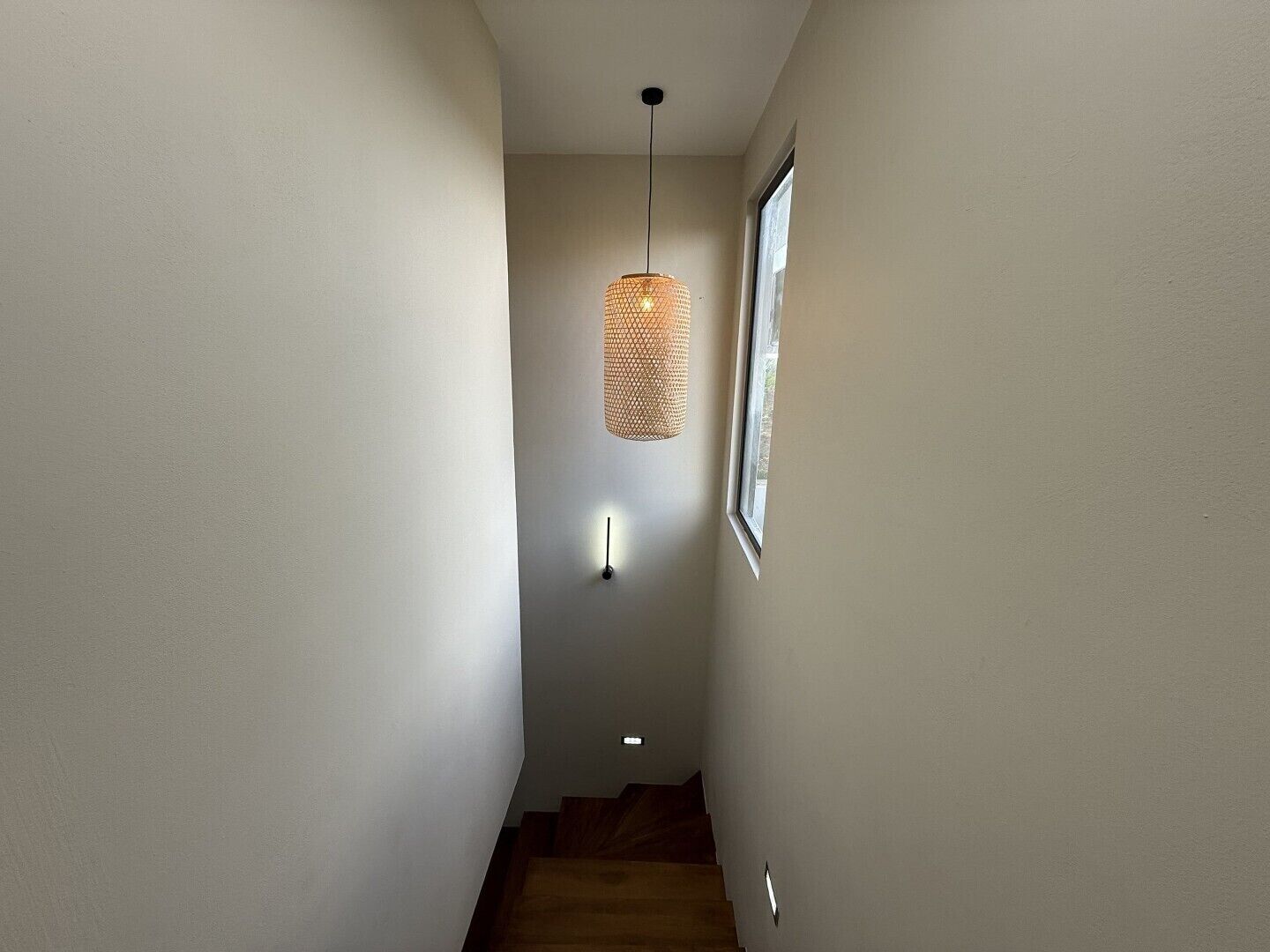 Ein schmaler Flur mit einer Holztreppe, einer strukturierten Pendelleuchte und einer Wandleuchte.