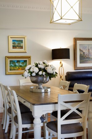 A dining area with a wooden table, white chairs, a flower vase, framed paintings, a black lamp, and a blue sofa.