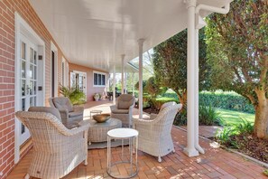 A covered patio with wicker furniture, a round table, and a brick building with a garden view.