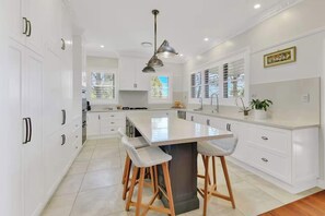 Une cuisine moderne avec des meubles blancs, un îlot central doté de tabourets, un luminaire suspendu et une fenêtre avec des stores.