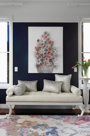 Ein Zimmer mit dunkelblauer Akzentwand, einem weißen Sofa mit gemusterten Kissen, einem floralen Kunstwerk und einer Vase mit Blumen auf einem Sockel.