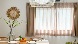 Ein Essbereich mit einem Holztisch, Stühlen, einer gewebten Pendelleuchte, einer Wanduhr und einem Fenster mit Vorhängen.