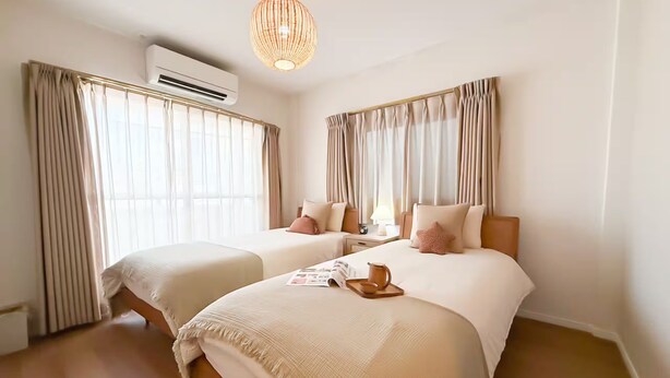 Ein Hotelzimmer mit zwei Betten, einem großen Fenster mit Vorhängen, einer Klimaanlage, einer Hängelampe und einem Nachttisch mit Lampe.