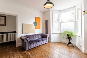 A room with a tufted sofa, a radiator, a window with a view, and a hanging light fixture. - 2 Bed Garden Retreat by Victoria Park (London)