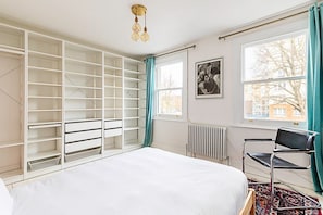 A bedroom with a bed, a window with curtains, a radiator, a chair, and a wall-mounted shelf unit. - 2 Bed Garden Retreat by Victoria Park (London)
