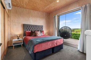 Ein Schlafzimmer mit Bett, Nachttisch, Lampe und Blick auf Grün hinter einem großen Fenster.